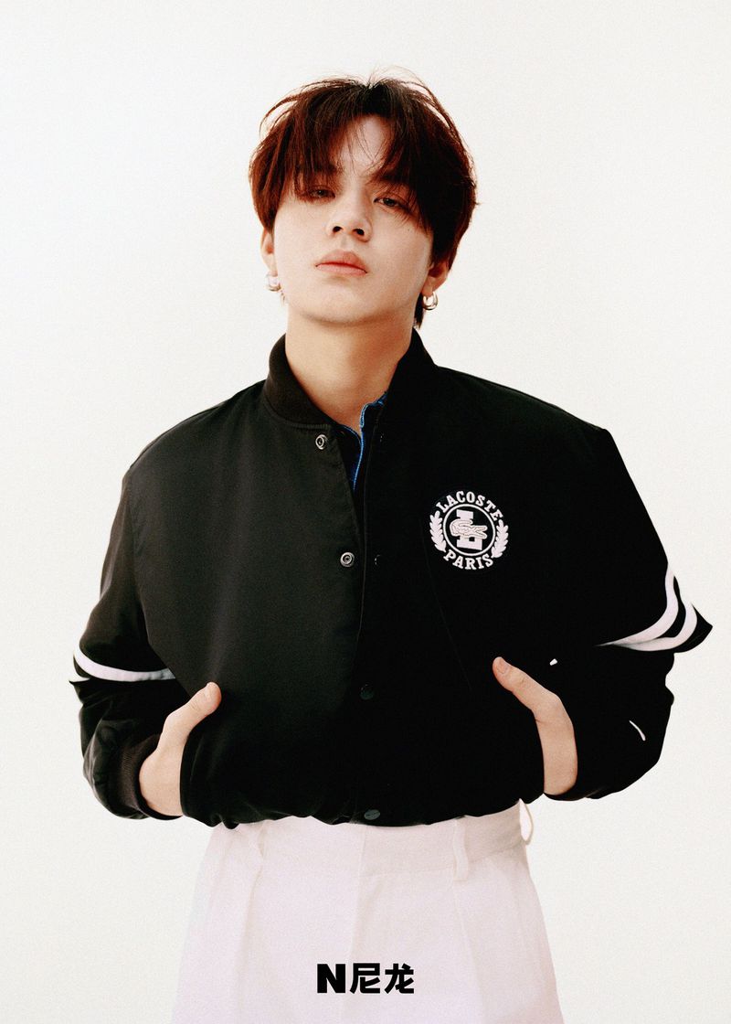 Portrait of a young adult wearing a Lacoste jacket and white pants against a white background, photographed in a studio with even lighting.