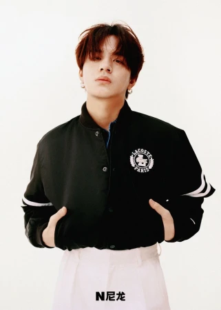 Portrait of a young adult wearing a Lacoste jacket and white pants against a white background, photographed in a studio with even lighting.