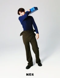 A man wearing a blue long-sleeve top and dark trousers holds a blue case over his shoulder against a plain white background.