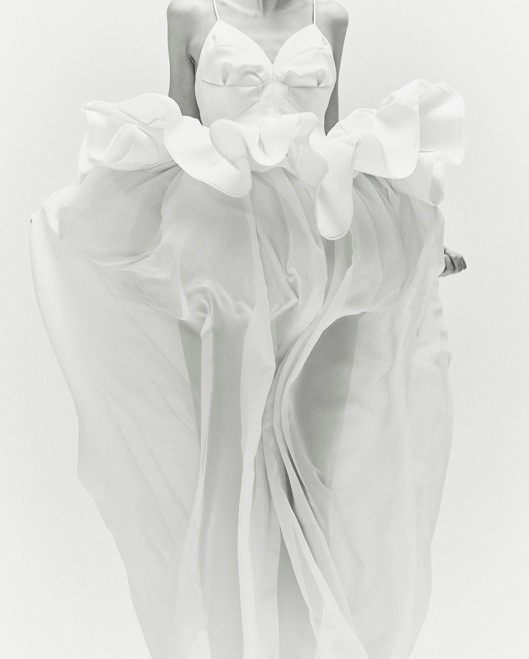 Elegant woman in a flowing white gown with ruffles, captured in a bright, monochrome studio setting, showcasing fashion and style.