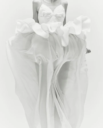 Elegant woman in a flowing white gown with ruffles, captured in a bright, monochrome studio setting, showcasing fashion and style.
