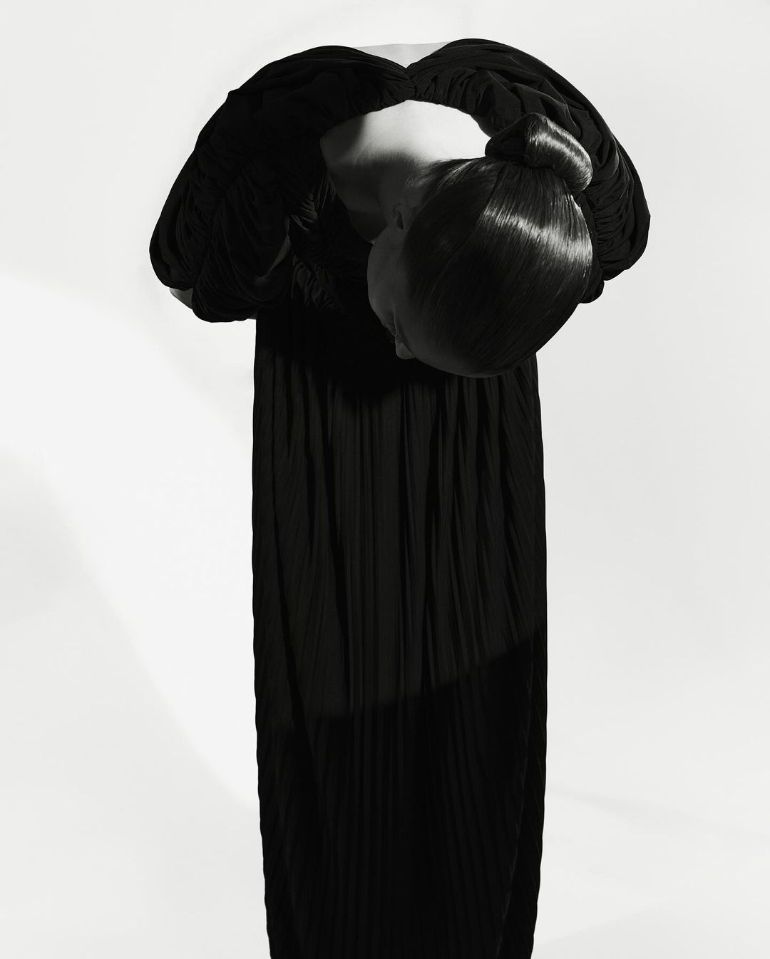 Model bowing forward in a long black dress with hair styled in a sleek updo against a white seamless background