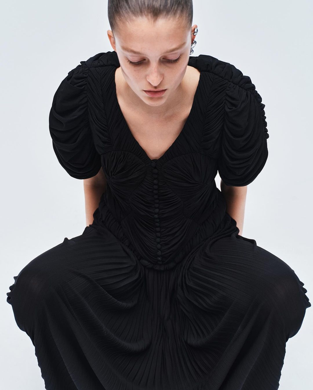 A woman in a black, textured midi dress with a V-neck and puffed sleeves poses against a white background in a studio shot.