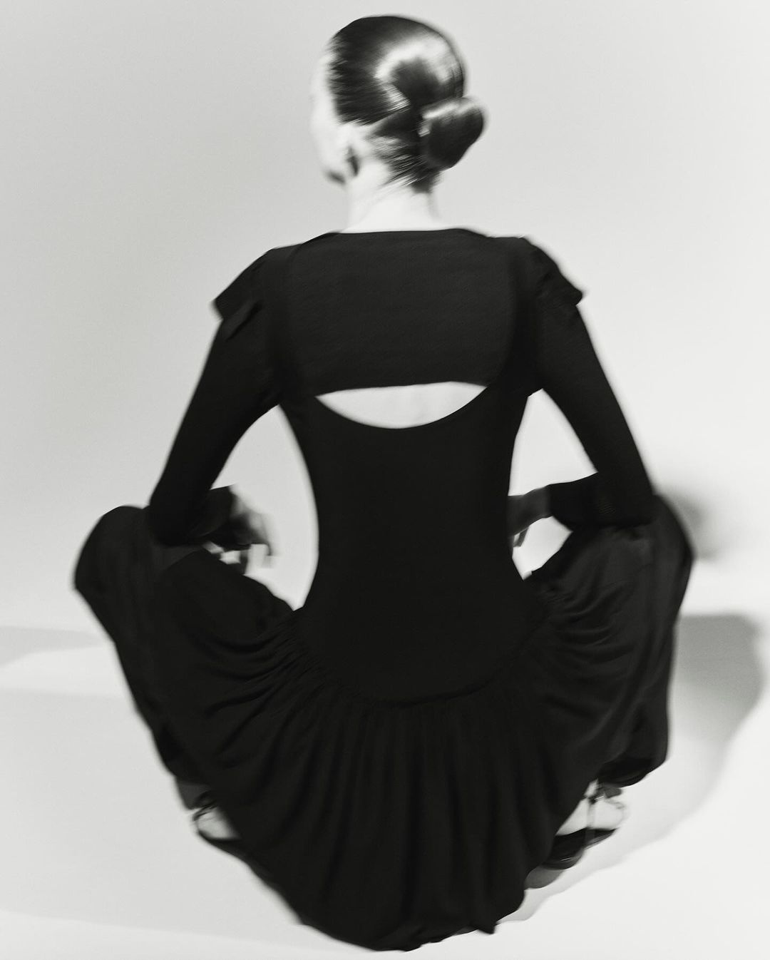 Model seen from behind wearing black top and trousers sitting against a light background