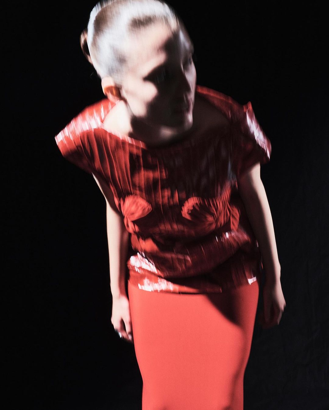 A woman with a blurred motion effect, wearing a red top and pencil skirt, poses in front of a dark, shadowy background for a fashion shot.