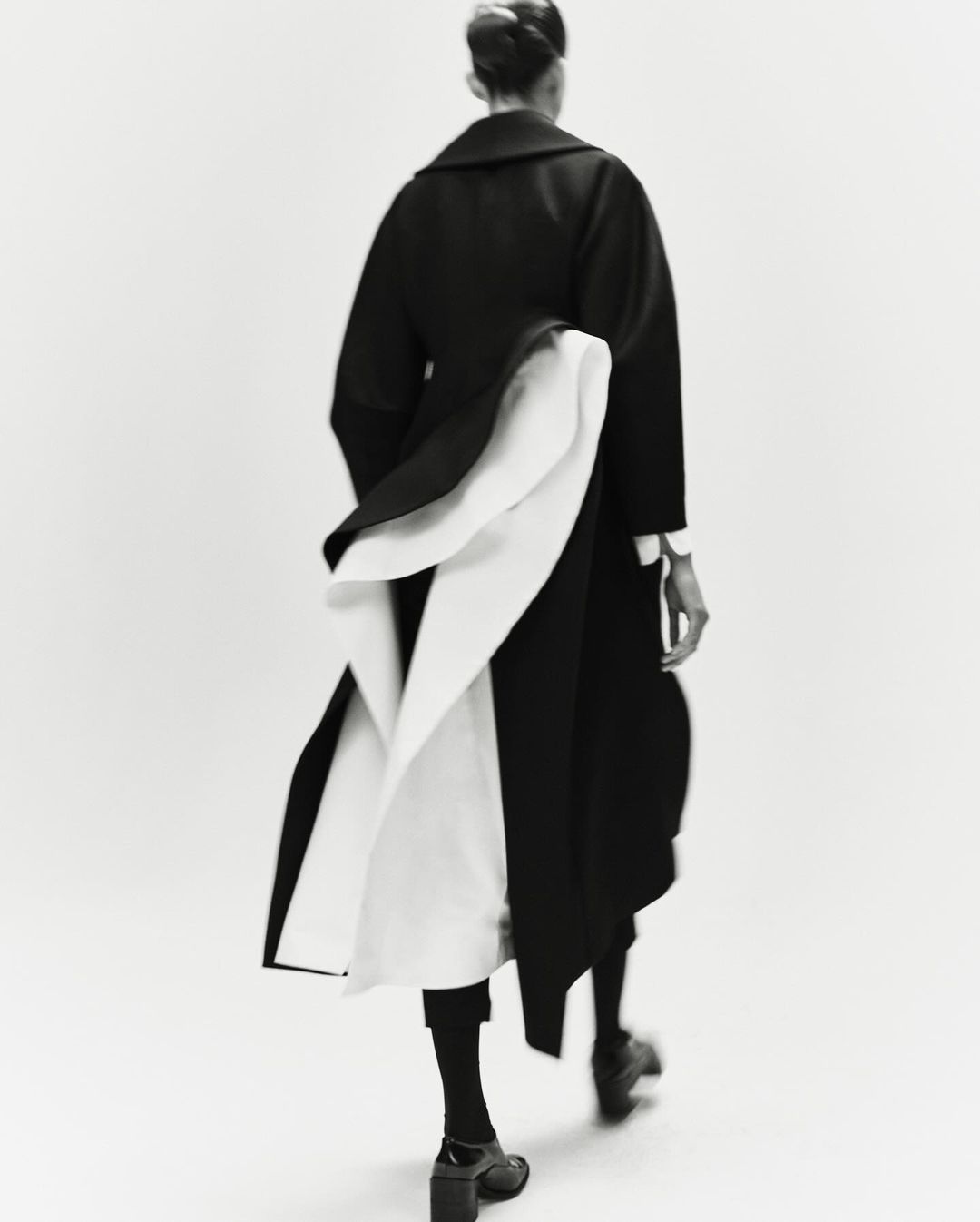 Back view of a fashion model in a black and white coat walking in a photo studio with a neutral background, editorial fashion photography.