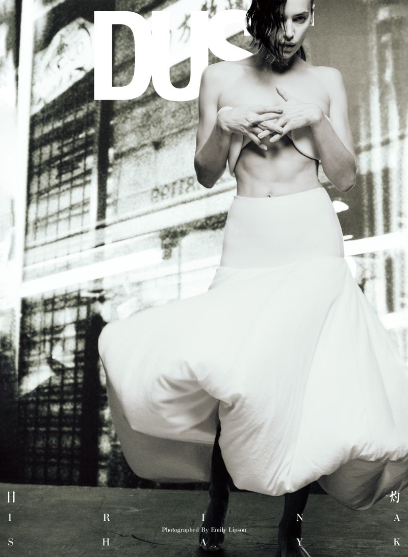 Fashion editorial shot of an adult female model wearing a white dress, posing in front of an abstract urban building backdrop.