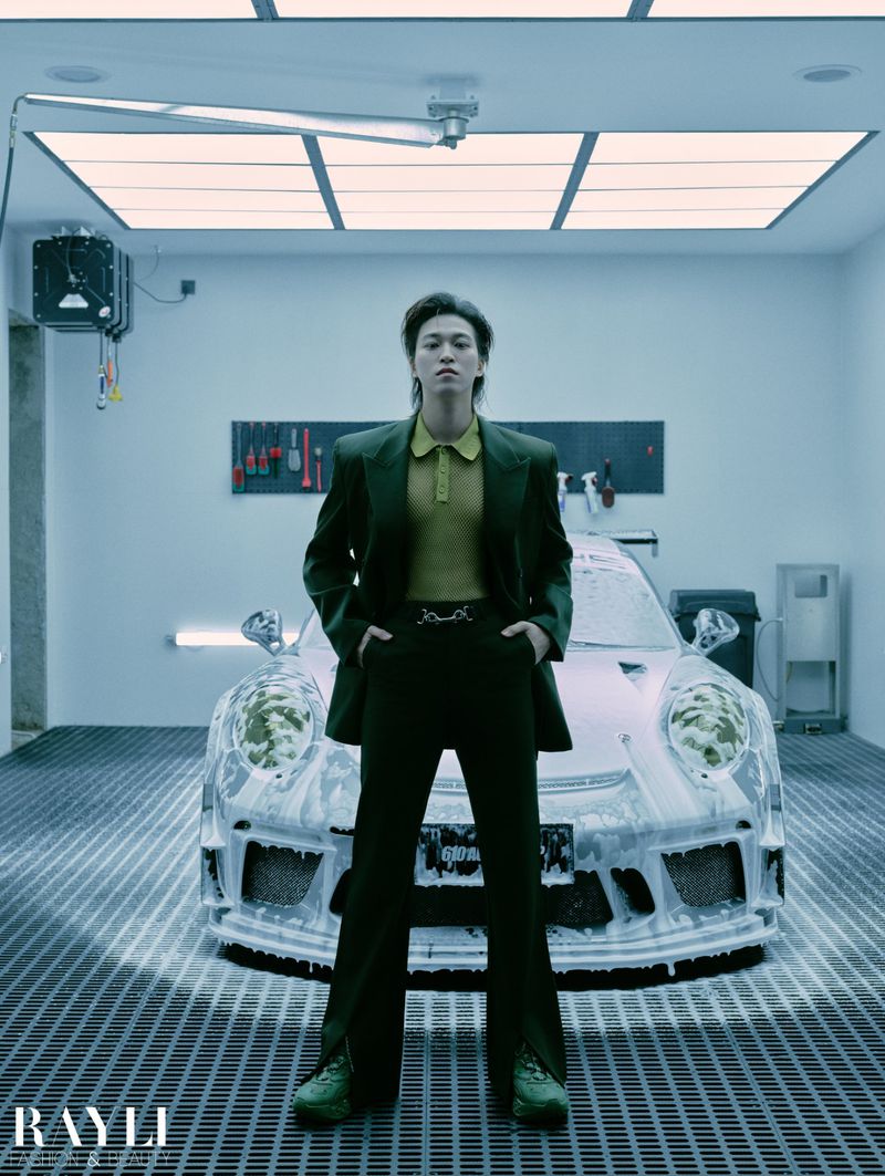 Man in dark suit poses in front of foamy Porsche inside garage with industrial lighting and tools in organized wall racks.