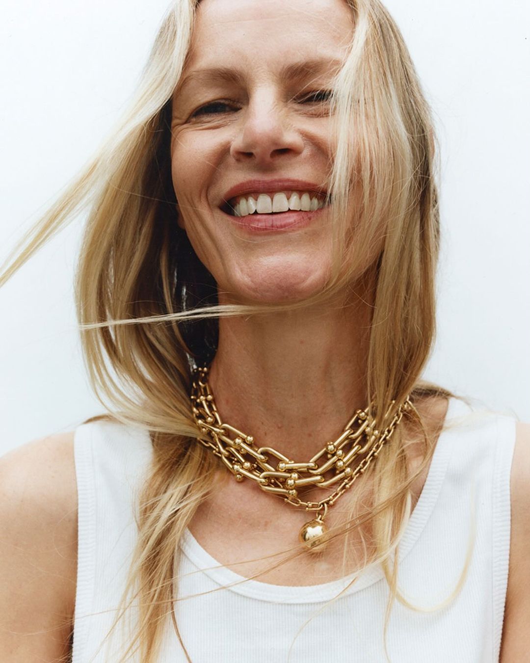 Smiling blonde woman poses indoors wearing a gold-tone chain-link necklace and white ribbed top, against a white backdrop.