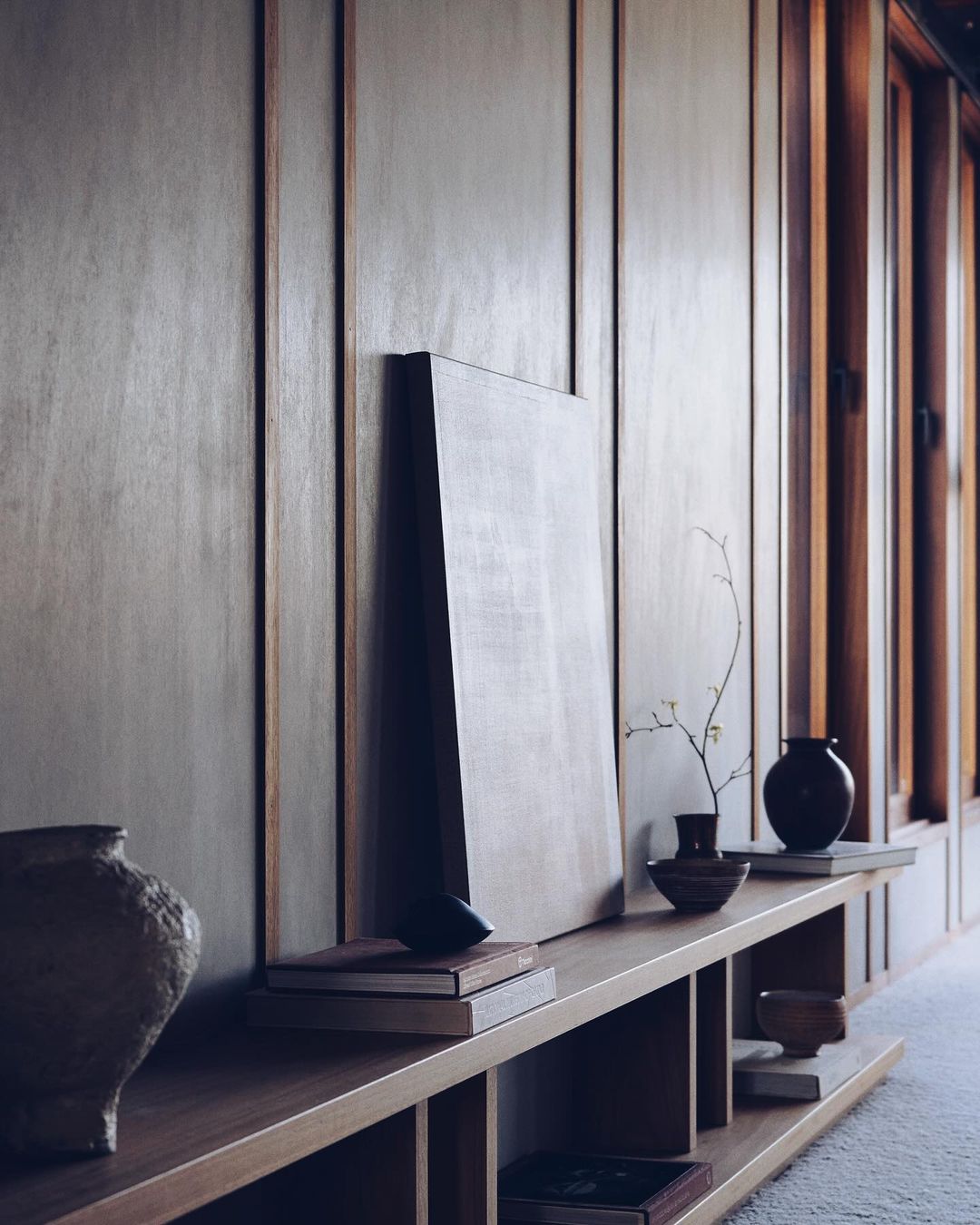 Minimalist interior with wooden shelving, vases, books, and a blank canvas against a paneled wall, conveying a serene and artistic atmosphere.