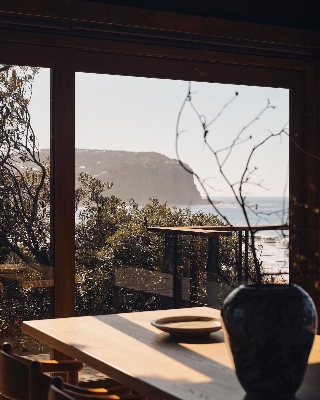 Interior view of a wooden dining table with chairs, a vase with branches, and a plate, seen through a window with a coastal view.