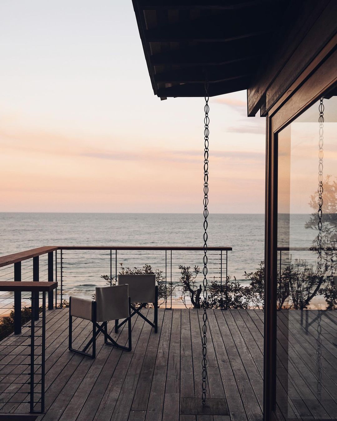 View from a wooden deck with chairs overlooking the ocean at sunset, with pink and orange sky reflected on the water.