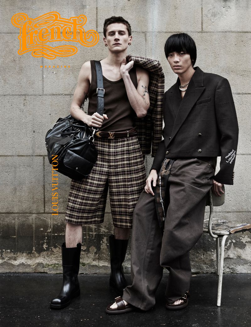 Two male fashion models pose in an urban setting, wearing contemporary clothing and accessories, with a concrete wall as the backdrop.