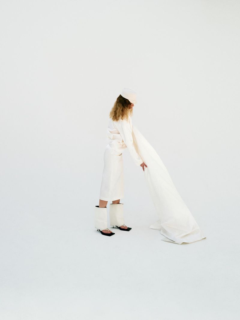 Fashion shot of a woman in an all-white outfit with a beret and fur-trimmed boots holding a flowing white fabric in a white photo studio.
