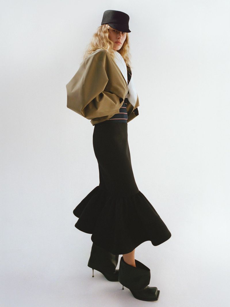 A woman poses in a khaki jacket, black skirt, hat, and high-heeled boots against a white backdrop in a fashion-editorial setting.