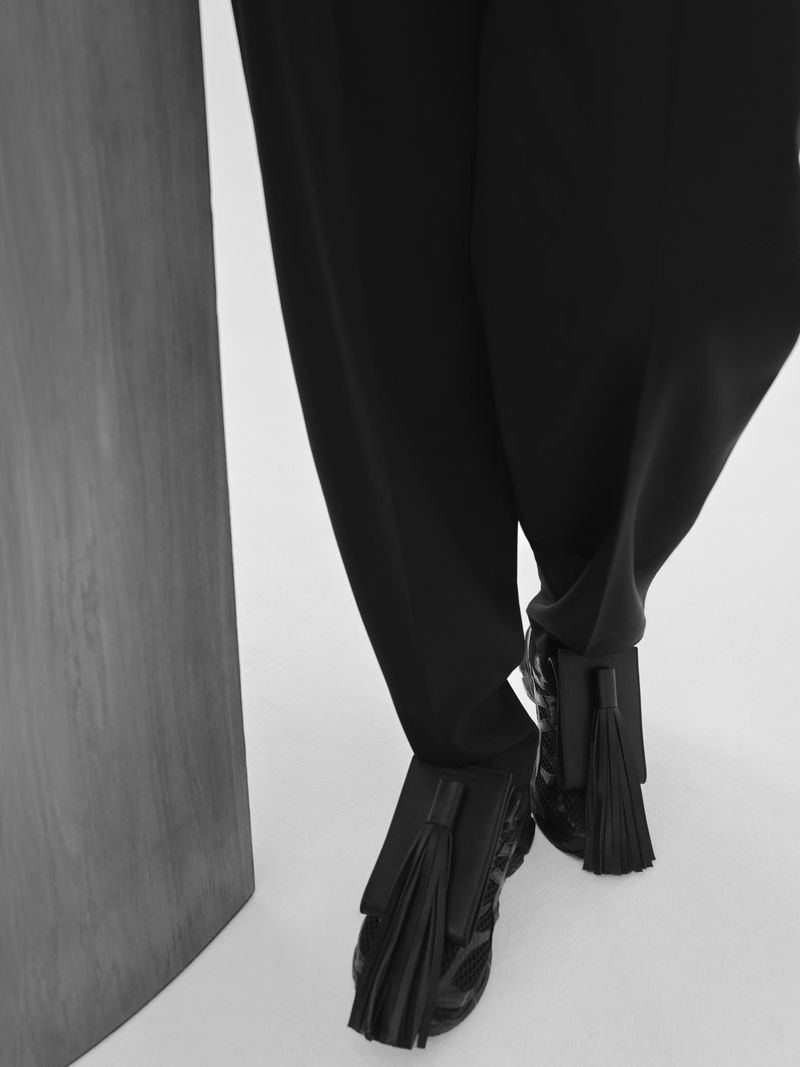 Close-up of person wearing black dress pants and tassel loafers standing in a minimalist interior setting with a light background.