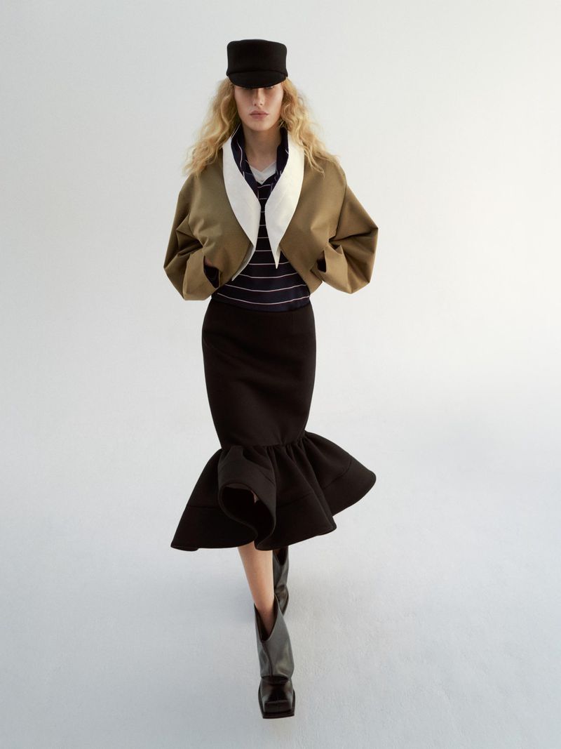 A woman poses in a khaki jacket, striped top, black pencil skirt with flared hem, boots, and a hat against a white backdrop in a fashion editorial.