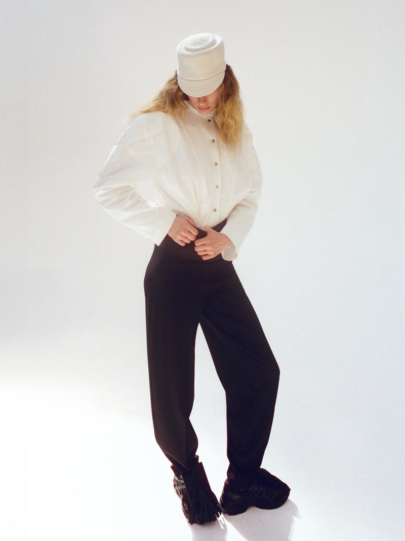 A woman in a white long-sleeved blouse, black pants, and black chunky shoes poses against a white backdrop wearing a white hat.