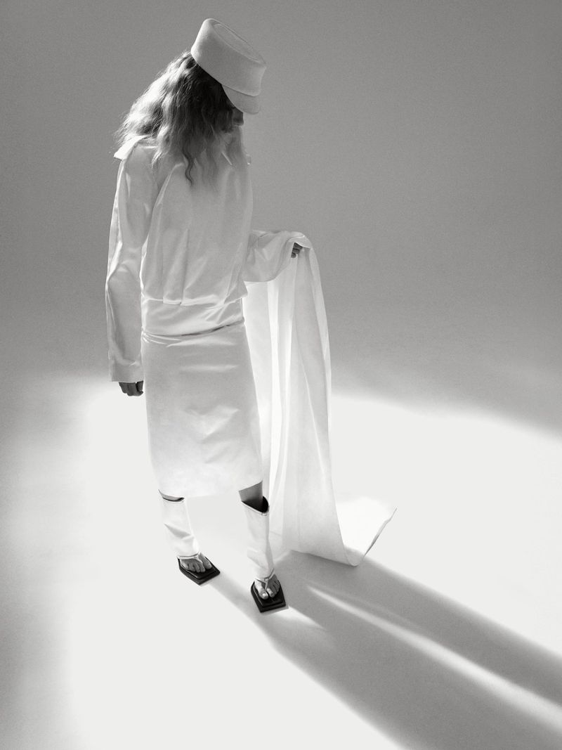 Monochromatic studio shot of androgynous person in white uniform and hat holding a white cloth, shot from a high angle.