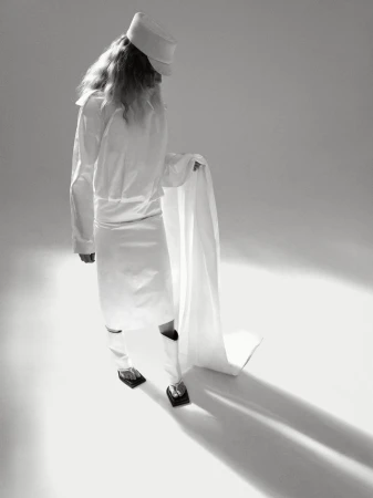 Monochromatic studio shot of androgynous person in white uniform and hat holding a white cloth, shot from a high angle.