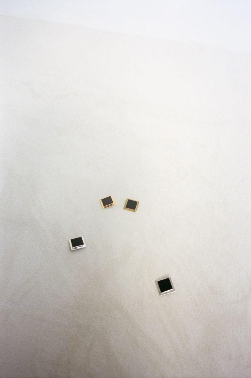 Four square stud earrings, two with silver trim and two with gold trim, arranged on a white surface with subtle textural details.