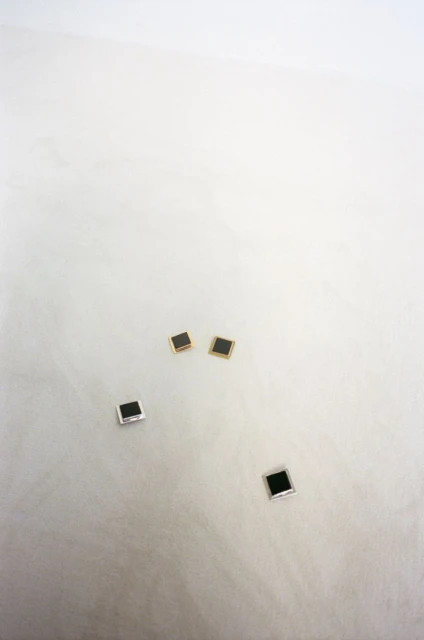 Four square stud earrings, two with silver trim and two with gold trim, arranged on a white surface with subtle textural details.