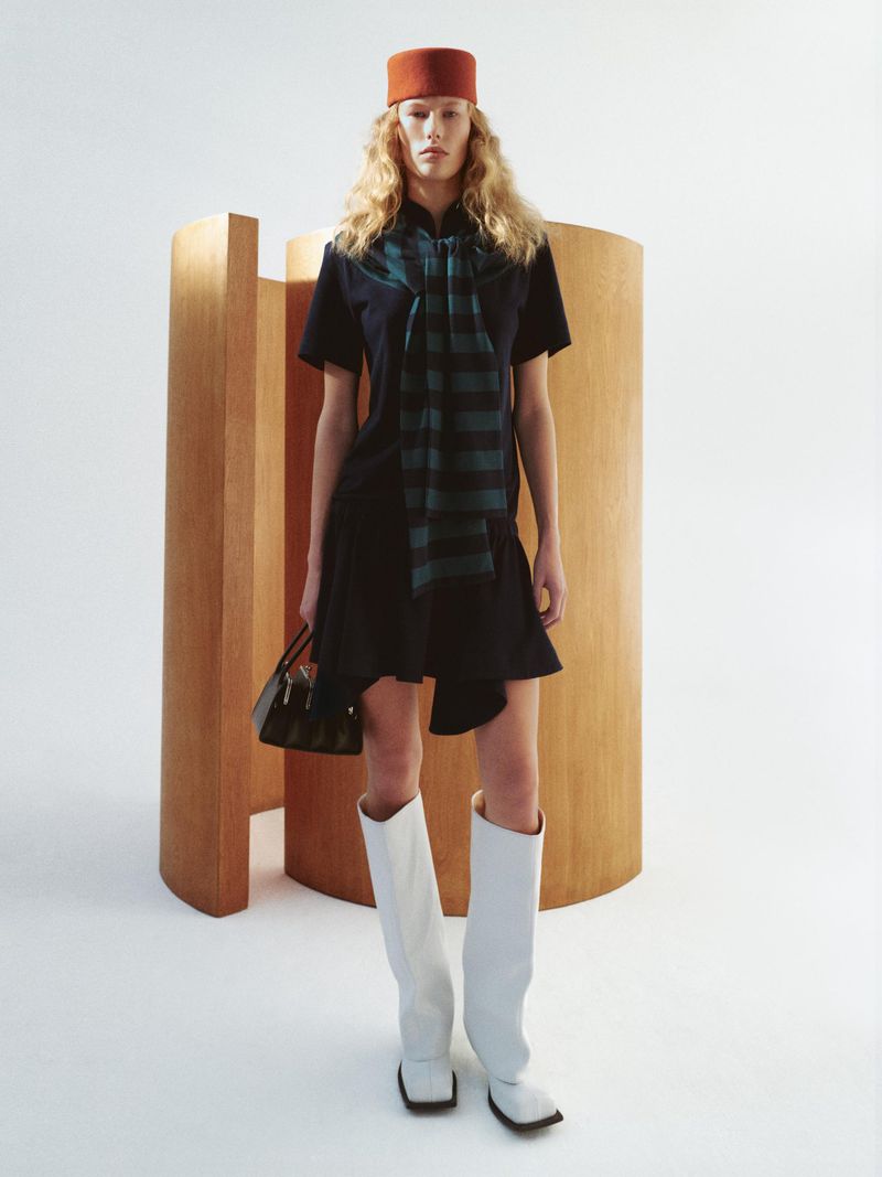 A solo adult female model poses in a dark blue mini dress with white knee-high boots, holding a purse and wearing a red hat indoors.