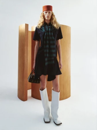 A solo adult female model poses in a dark blue mini dress with white knee-high boots, holding a purse and wearing a red hat indoors.