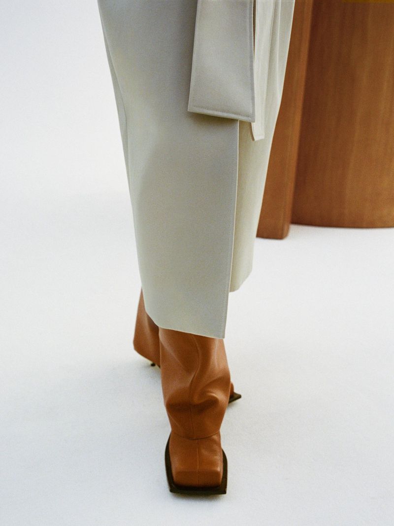 Model wears tan knee-high boots and long ivory skirt in a studio environment with a light wood accent table, fashion forward clothing.