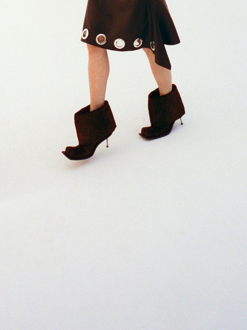 Fashionable woman wearing a brown mini skirt with silver eyelet detailing and unique brown heeled boots on a stark white surface.