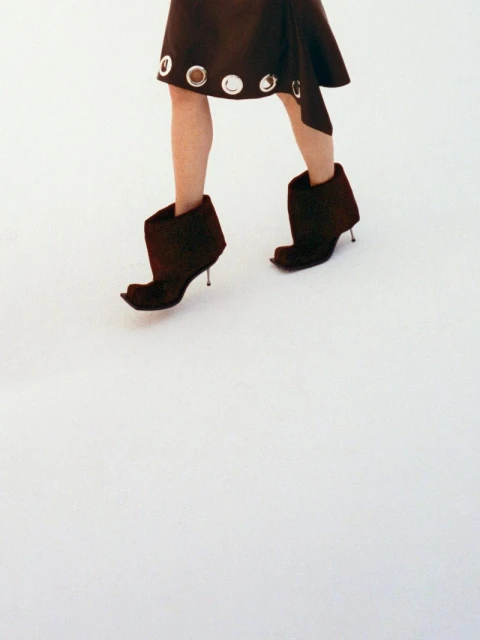 Fashionable woman wearing a brown mini skirt with silver eyelet detailing and unique brown heeled boots on a stark white surface.