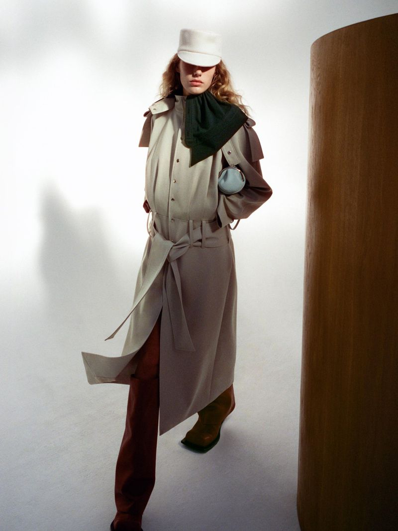 A model wears a beige belted trench coat and a white cap in a fashion forward studio portrait against a gradated white background.