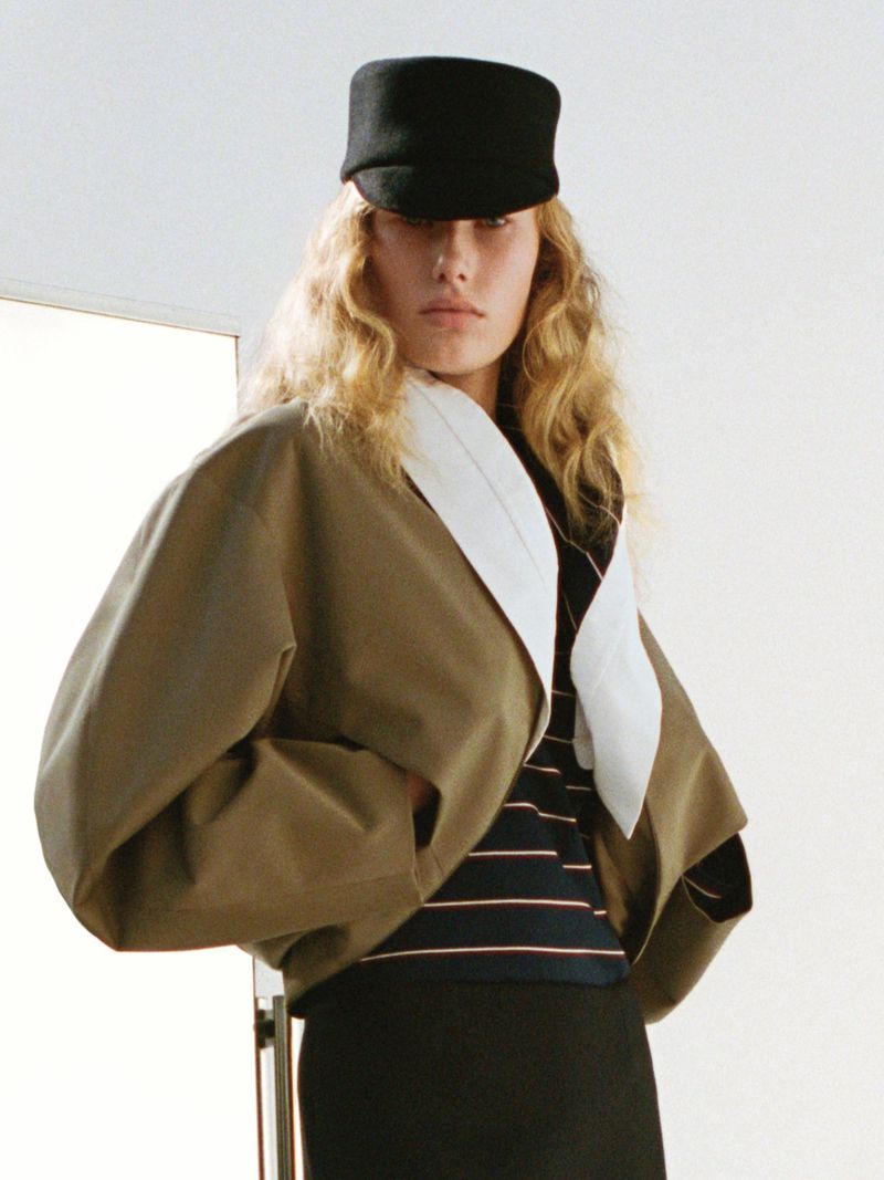 Woman wearing a black cap and earth-tone jacket poses in a studio with blonde hair and neutral makeup, fashion photography.