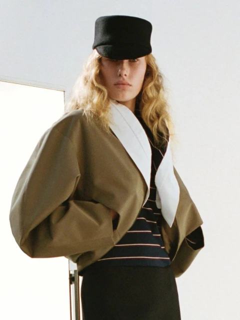 Woman wearing a black cap and earth-tone jacket poses in a studio with blonde hair and neutral makeup, fashion photography. 