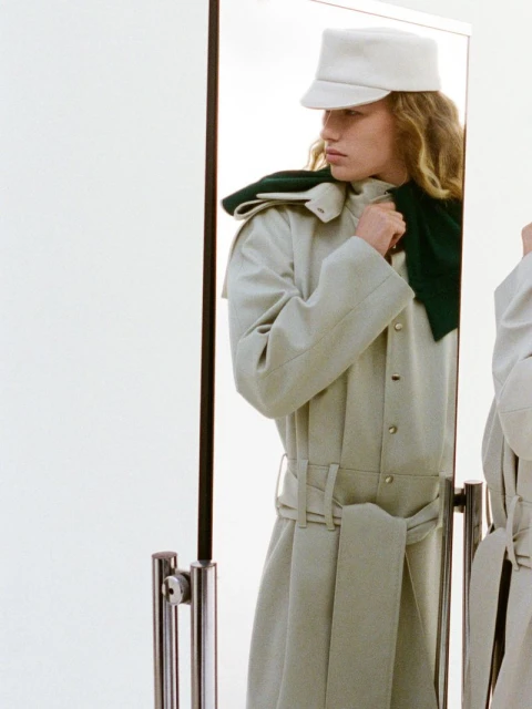 A model wearing a trench coat and cap poses in a photo studio, reflected in a mirror. The image has a bright, clean aesthetic.
