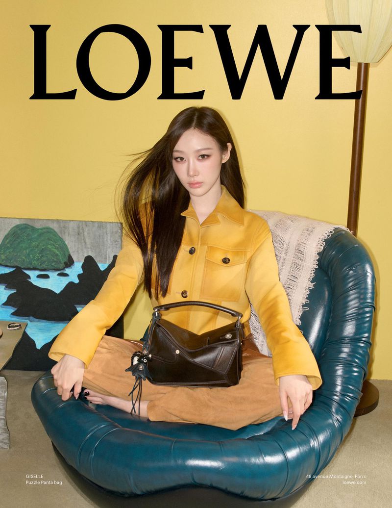 A woman sits in a teal chair with a black Loewe bag in a brightly lit interior against a yellow wall with the Loewe logo.