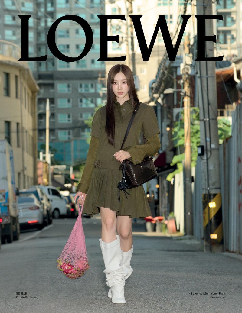 A woman models Loewe clothing in an urban setting, wearing a green dress, white boots, and carrying a Loewe bag and a pink mesh bag.