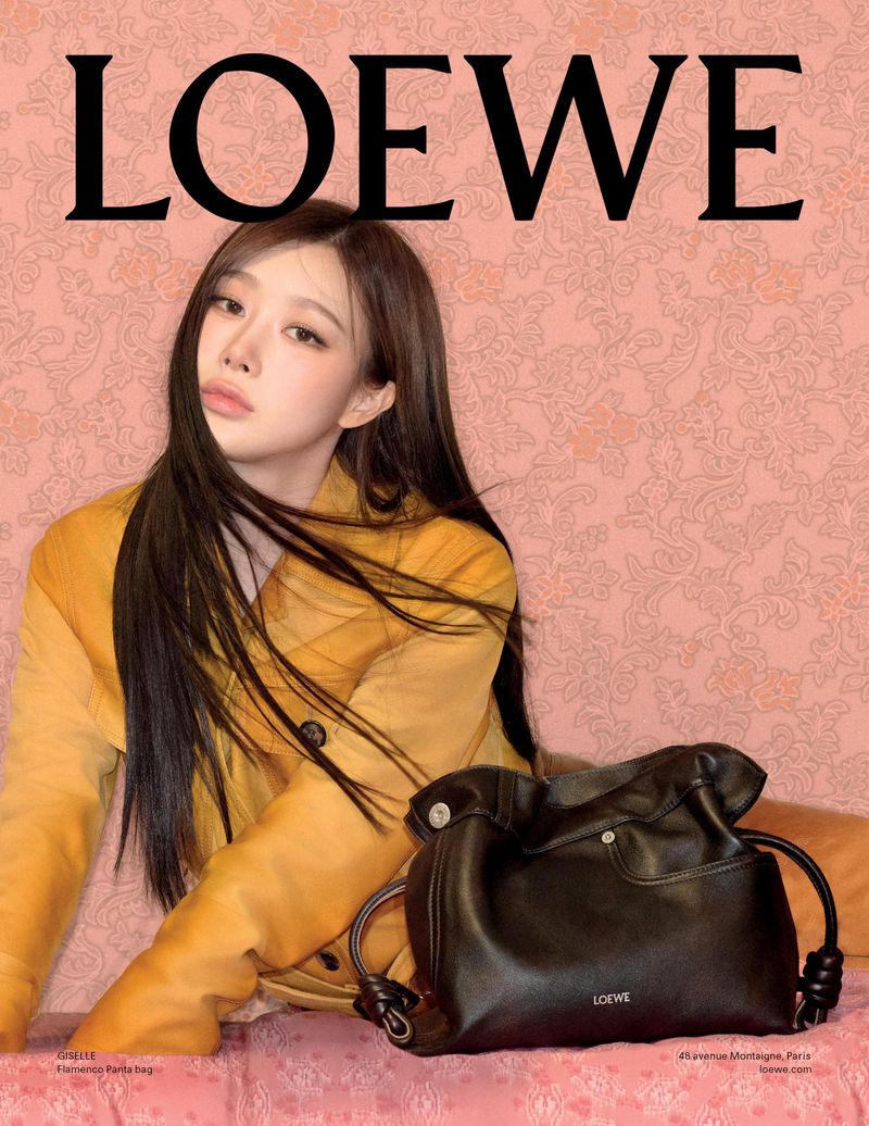 Woman with long dark hair wearing a yellow top next to a black leather handbag against a pink patterned backdrop with LOWE text above
