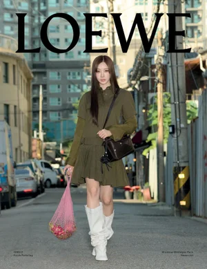 Giselle Uchinaga walking down a city street wearing an olive green dress and white boots carrying a pink string bag for Loewe