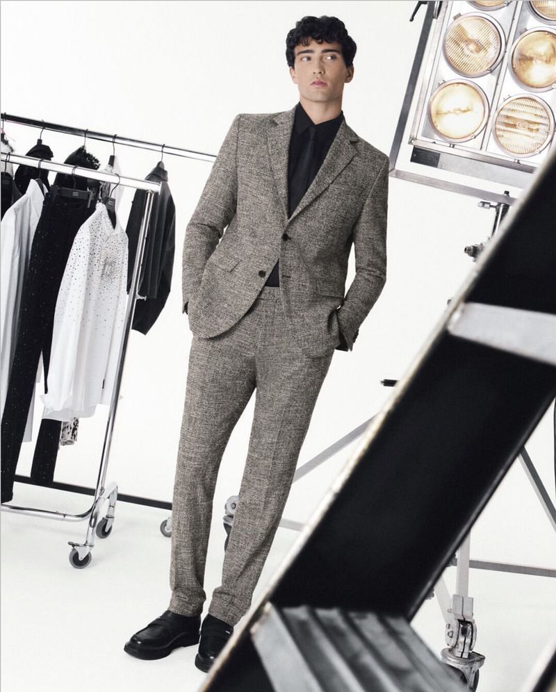 A male model poses in a grey patterned suit with a black shirt and black shoes in a studio with lights and clothing rack.