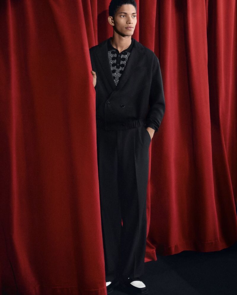 A young adult male model poses in a black suit, peering out from behind a red curtain in a studio fashion shot.