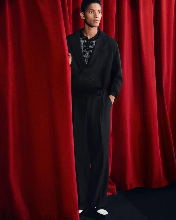 Male model wearing an all-black suit and patterned shirt standing between draped red velvet curtains.