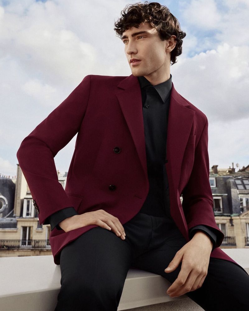 A handsome dark-haired man in a maroon jacket and black shirt poses on a rooftop against a cloudy sky and European buildings.