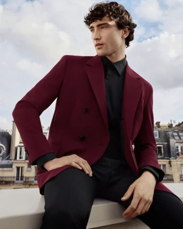 A handsome dark-haired man in a maroon jacket and black shirt poses on a rooftop against a cloudy sky and European buildings.