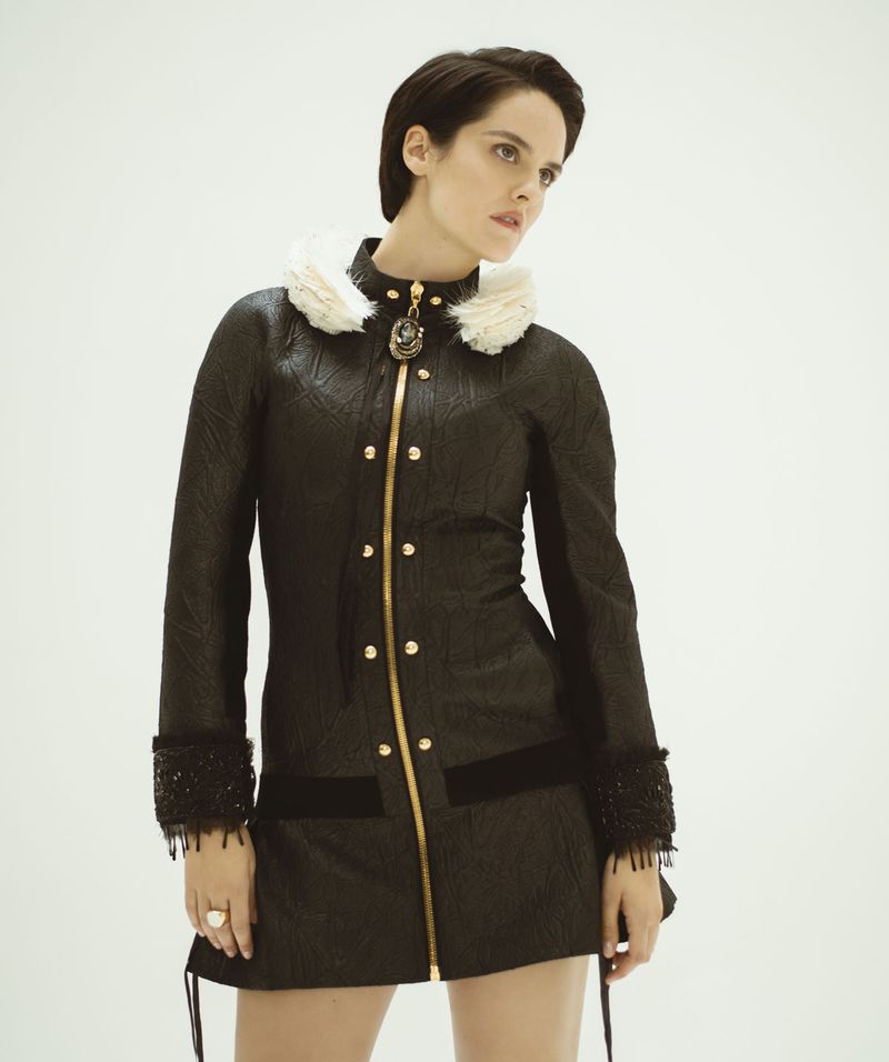 A woman in a dark textured coat with a fur collar poses in a studio. The coat has gold button and zipper details, and a decorative brooch.