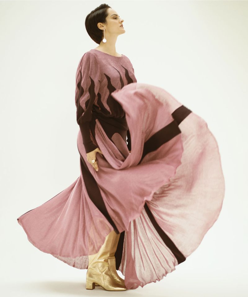 Woman in a flowing pink dress with brown accents and metallic gold boots poses in a studio with a white backdrop looking elegant.