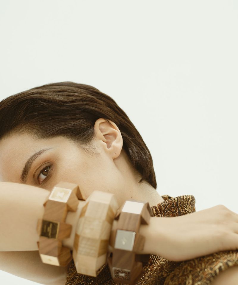 Close-up studio shot of an adult woman with short brown hair wearing multiple wood-tone blocky bracelets against a white backdrop.