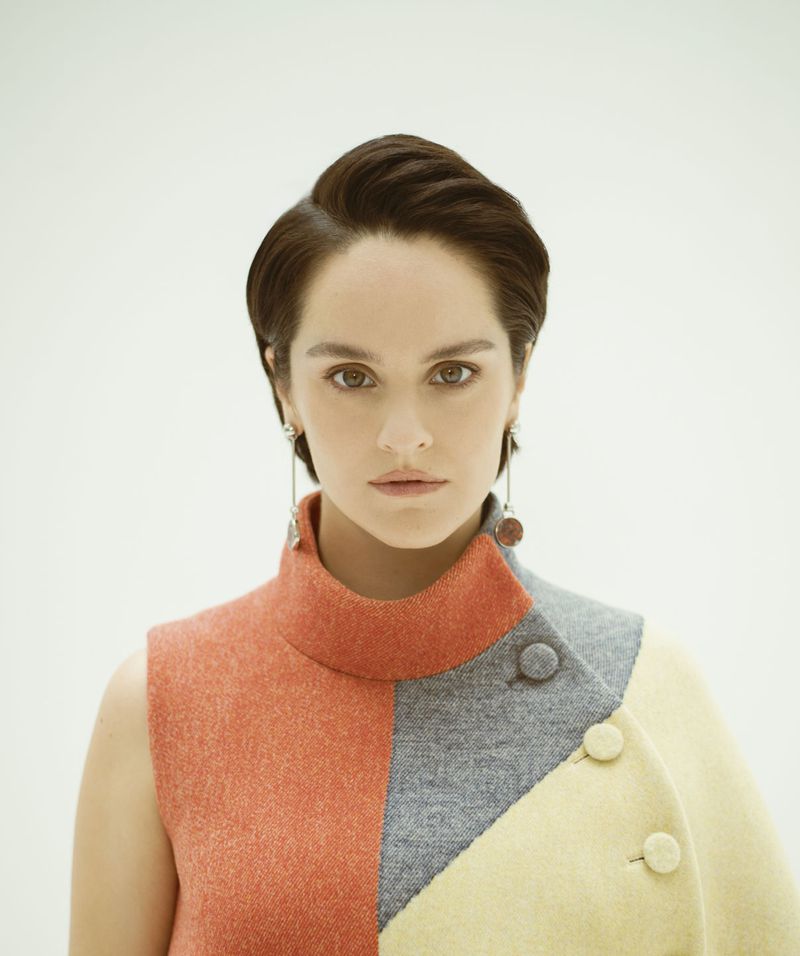 Portrait of a woman with short brunette hair wearing a color-blocked high-neck dress and dangling earrings against a neutral backdrop.