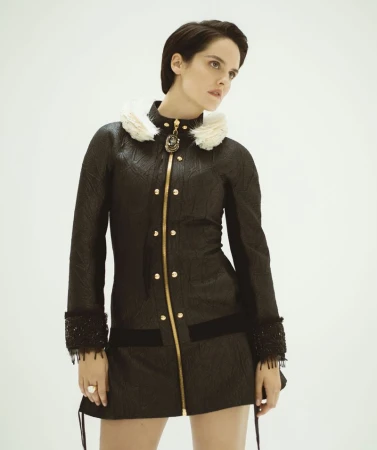 Portrait of actor Noemie Merlant wearing a black textured coat with a white feathery collar against a seamless white background.