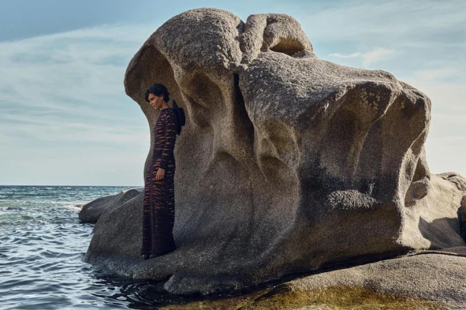 Model Polina Park posing inside a hollowed rock formation along a rocky shoreline wearing a sheer patterned dark maxi dress.